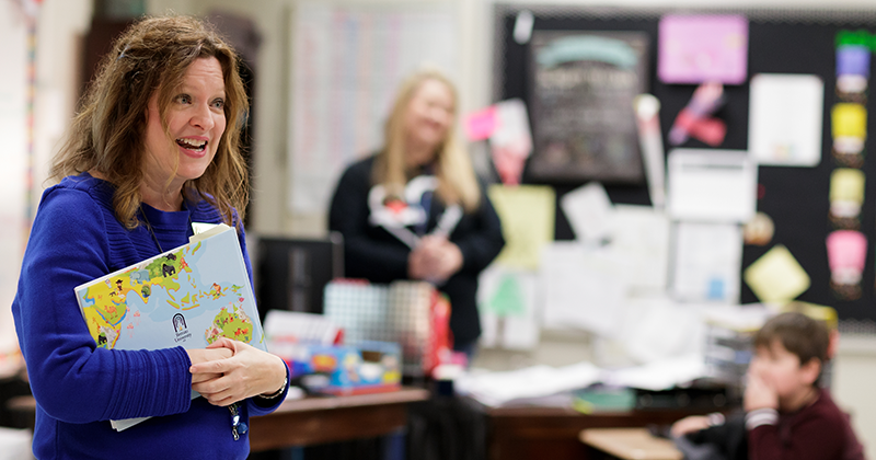smiling teacher holding a book in front of classroom