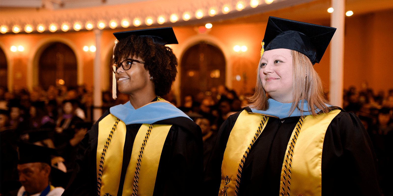 two women graduates as Education Specialists wearing black cap and gown with yellow ribbons