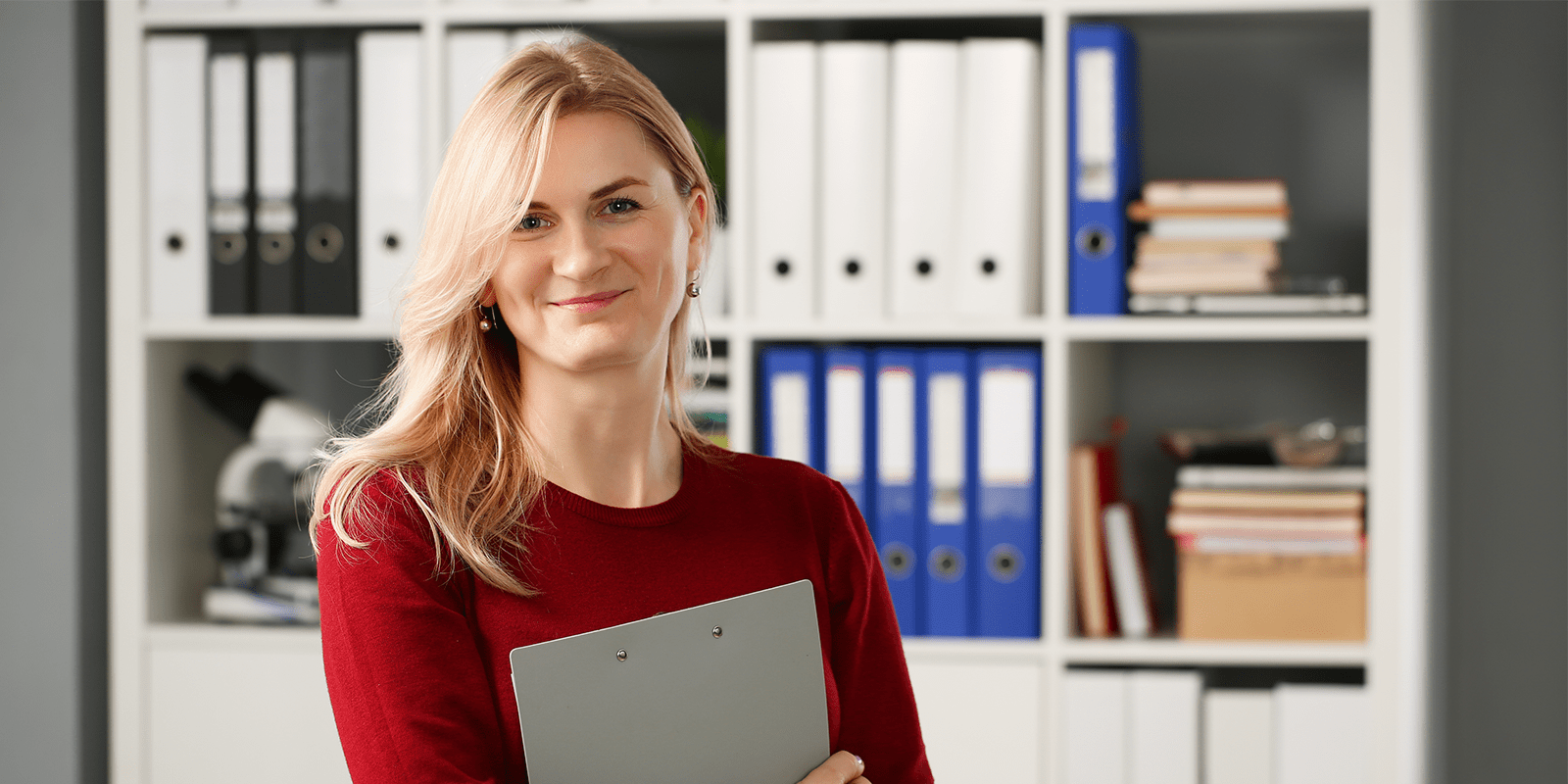 blonde woman in red shirt in front of shelf of binders