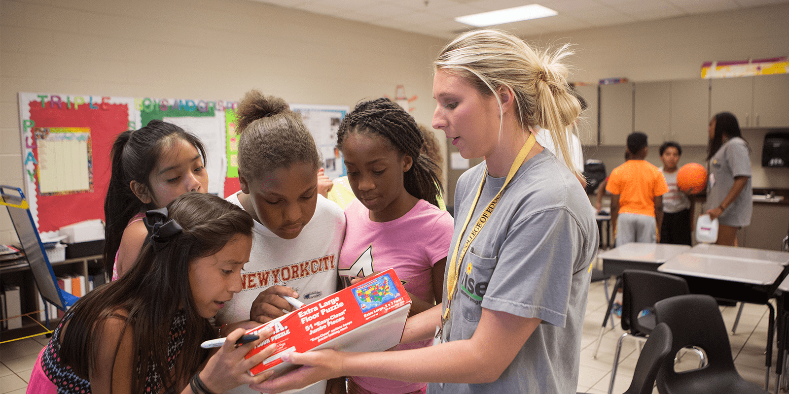 blonde middle grades education teacher holding puzzle in front of group of girls in classroom