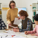 header-1920x1280-Teacher-Assisting-Students-During-Classroom-Group-Work Teacher helping diverse students review papers at classroom table