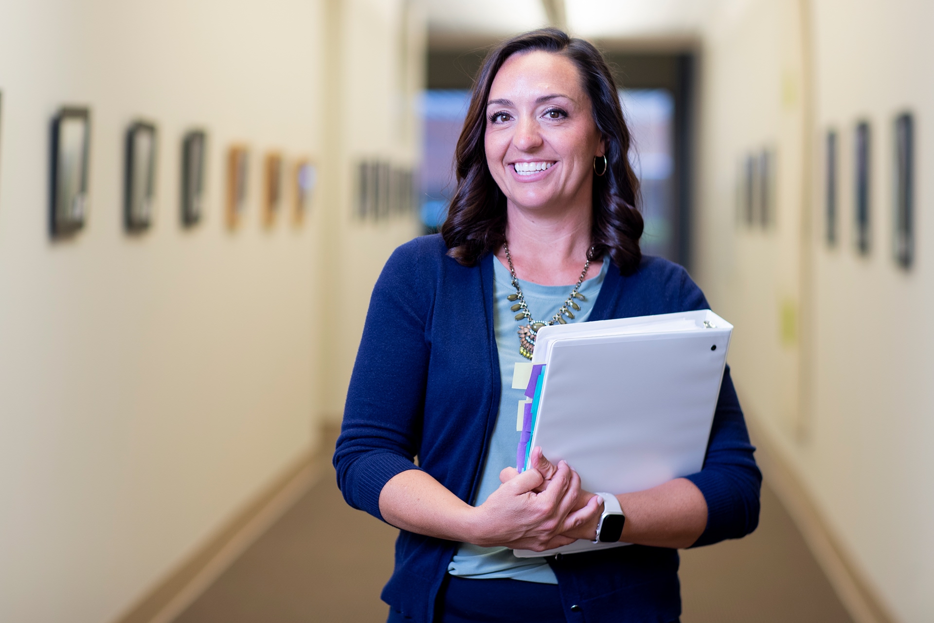 Smiling advisor in navy cardigan holding tabbed binder in office hallway